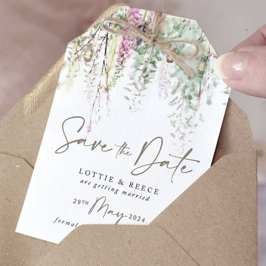 Rustic Save the Date cards cut into a tag shape and finished with a jute twine bow. The design features sage green foliage with whimsical flowrers and string lights. The typography is simple and modern with caligraphy style accents.